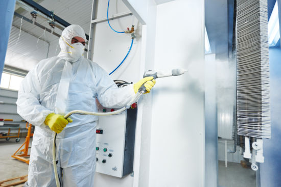 industrial metal coating. Worker man in protective suit with gas mask spraying powder to steel finished parts in painting chamber